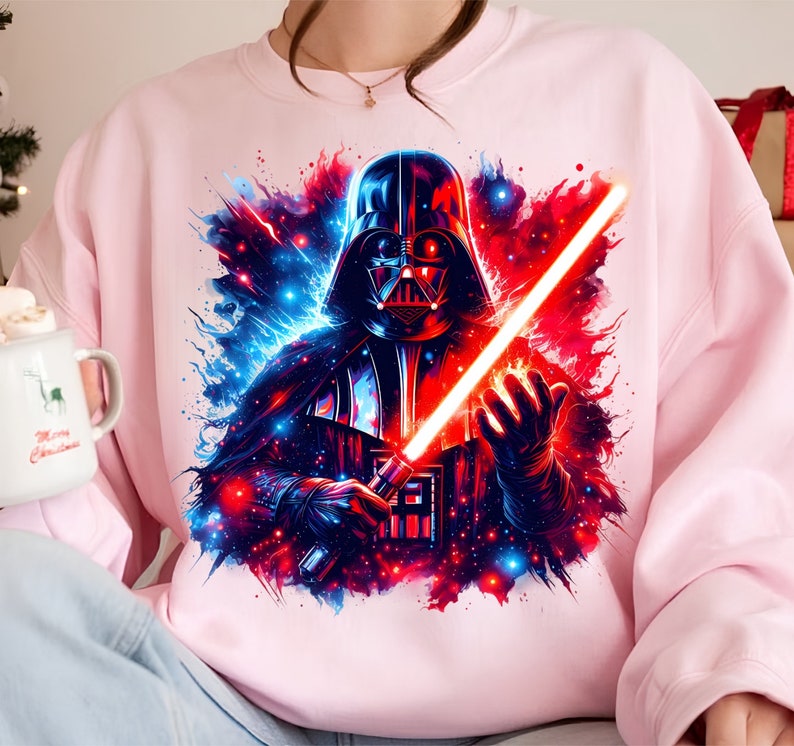 May include: A pink sweatshirt with a graphic print of Darth Vader holding a red lightsaber. The background is a galaxy with blue and red stars.