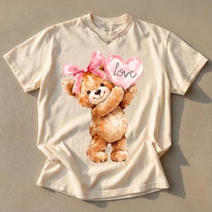 May include: Cream-colored t-shirt featuring a watercolor illustration of a teddy bear holding a pink heart with the word "Love" written on it. The bear has a pink bow and is smiling. The shirt has a distressed neckline.