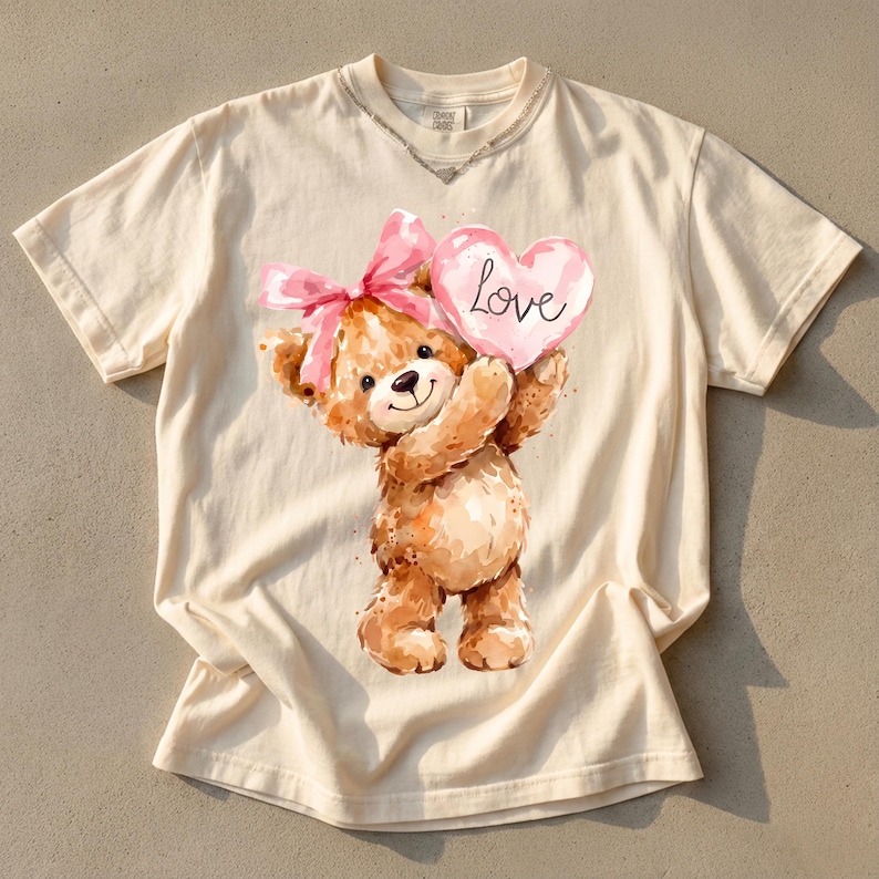 May include: Cream-colored t-shirt featuring a watercolor illustration of a teddy bear holding a pink heart with the word "Love" written on it. The bear has a pink bow and is smiling. The shirt has a distressed neckline.