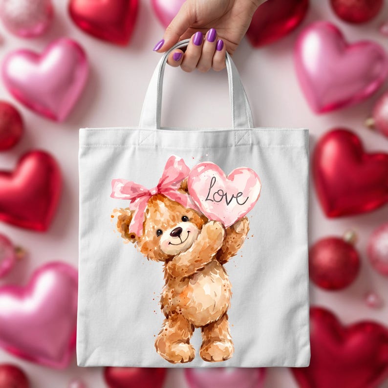May include: White tote bag featuring a watercolor illustration of a teddy bear holding a pink heart with the word "Love". The bear has a pink bow and is surrounded by pink and red heart-shaped balloons.