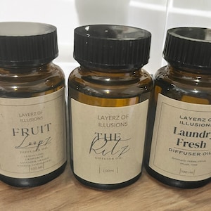 May include: Three brown glass diffuser oil bottles with black caps. Each bottle has a label with the text "LAYERZ OF ILLUSIONS". The labels read "Fruit Loopz", "The Ritz", and "Laundry Fresh". Each bottle contains 100 ml of oil.