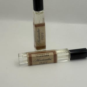 May include: Two small, rectangular perfume bottles with black lids. The bottles are clear, filled with a light-coloured liquid, and have labels that read "Layerz of Illusions Marshmallow Puff."