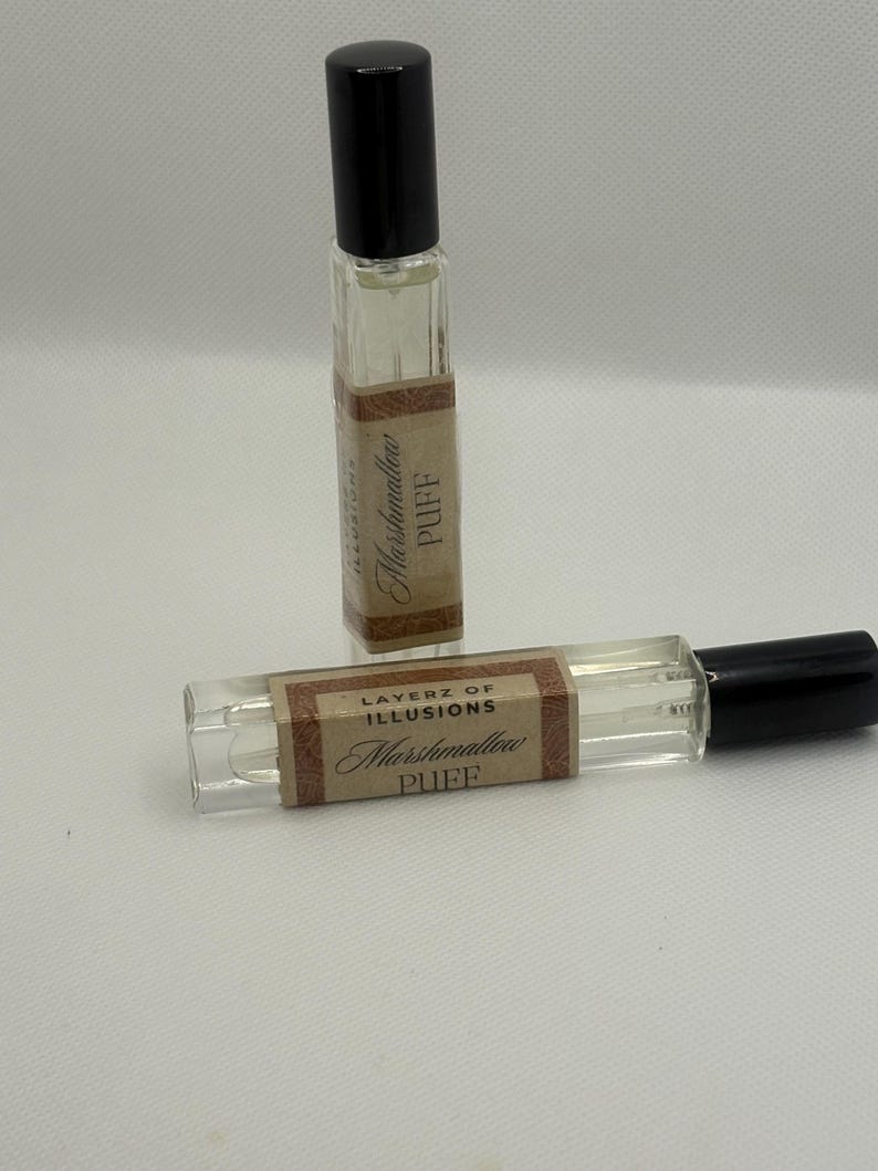 May include: Two small, rectangular perfume bottles with black lids. The bottles are clear, filled with a light-coloured liquid, and have labels that read "Layerz of Illusions Marshmallow Puff."