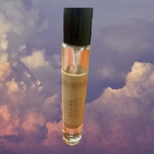 Puede incluir: Un frasco transparente de vidrio con aplicador de bola, con una tapa negra y una etiqueta marr&oacute;n que dice "DIVINE CLOUDS". El frasco est&aacute; fotografiado sobre un fondo de nubes blancas y rosadas esponjosas.