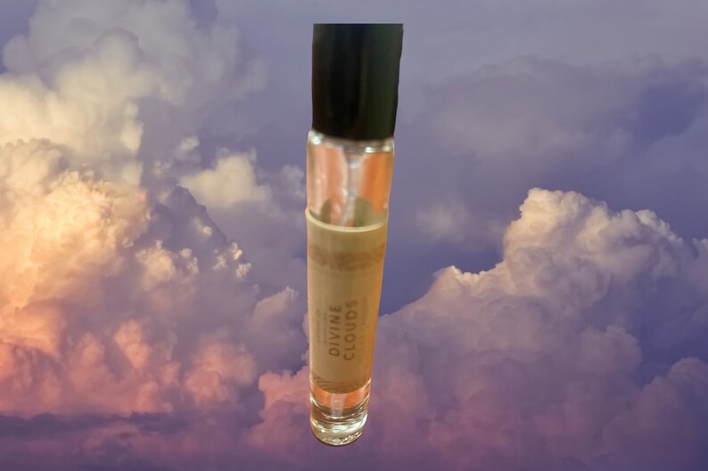 Puede incluir: Un frasco transparente de vidrio con aplicador de bola, con una tapa negra y una etiqueta marr&oacute;n que dice "DIVINE CLOUDS". El frasco est&aacute; fotografiado sobre un fondo de nubes blancas y rosadas esponjosas.
