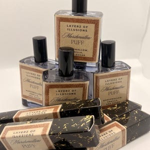 May include: A selection of Layerz of Illusions Marshmallow Puff perfume bottles and boxes. The bottles are clear with black caps and labels. The boxes are black with gold flecks and matching labels. The labels read "Marshmallow Puff".