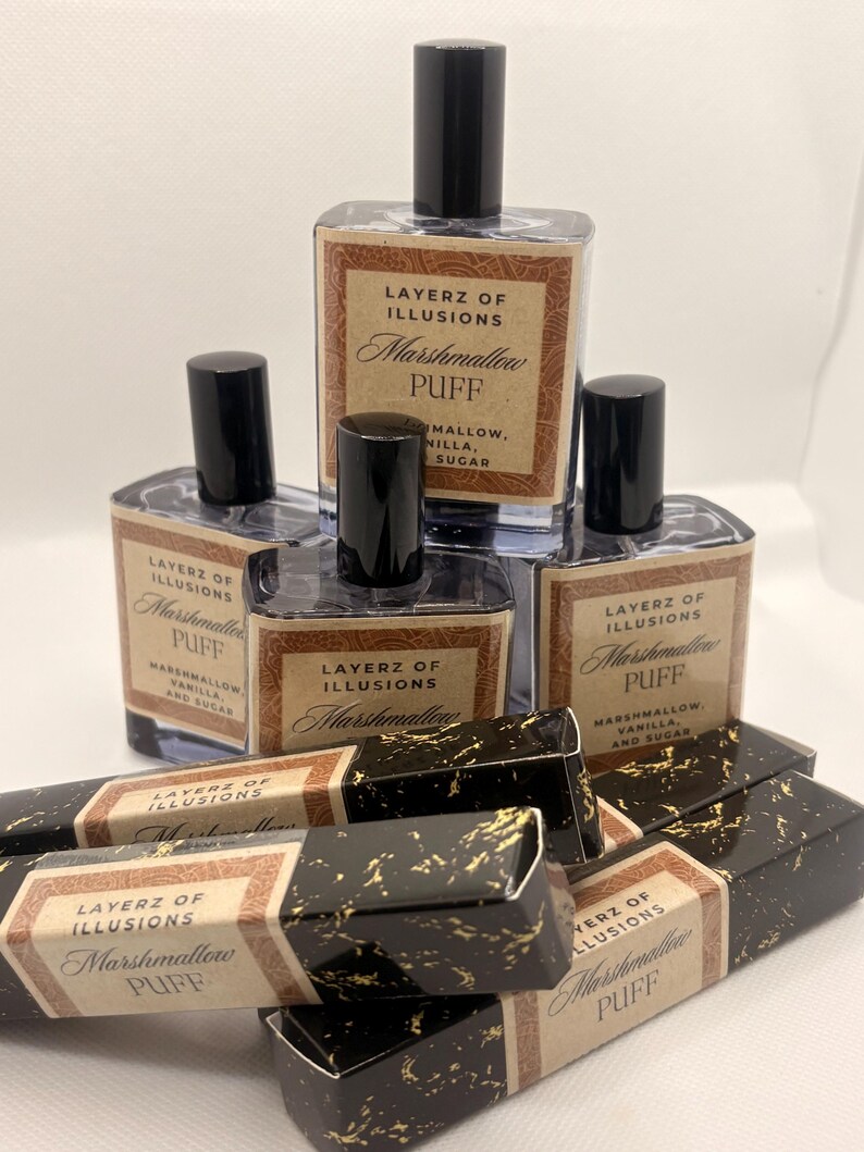 May include: A selection of Layerz of Illusions Marshmallow Puff perfume bottles and boxes. The bottles are clear with black caps and labels. The boxes are black with gold flecks and matching labels. The labels read "Marshmallow Puff".
