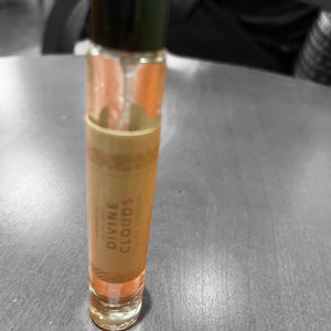 Puede incluir: Una botella de vidrio transparente con una tapa negra y una etiqueta que dice "DIVINE CLOUDS". La botella est&aacute; llena de un l&iacute;quido naranja claro.
