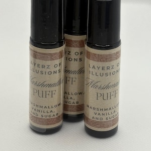 May include: Three roll-on bottles with black lids and labels reading "LAYERZ OF ILLUSIONS Marshmallow PUFF" with ingredients. The bottles are cylindrical and rest on a white surface.