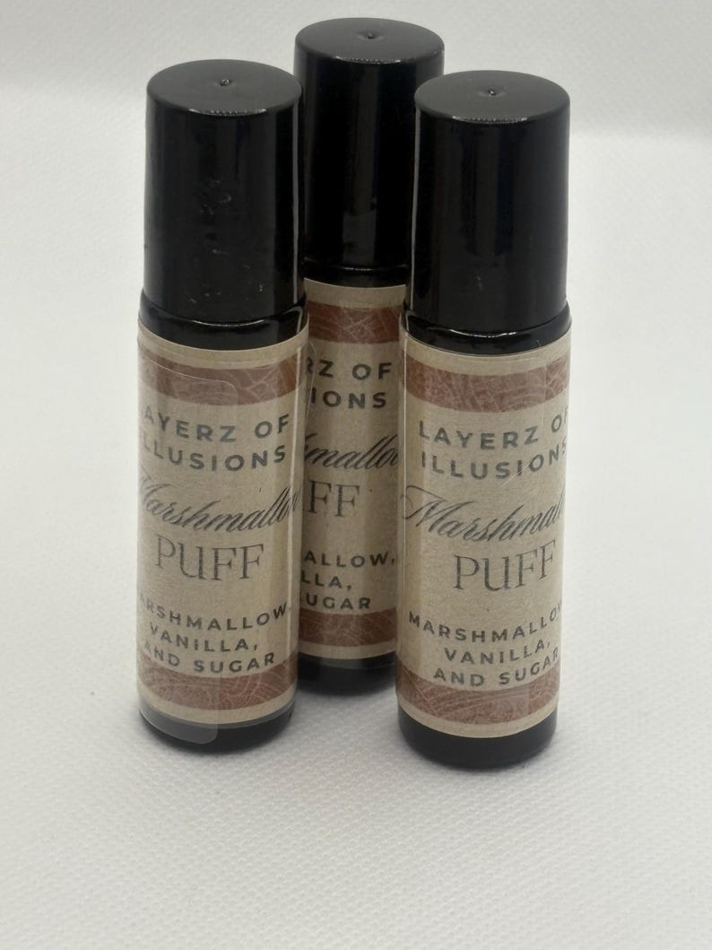 May include: Three roll-on bottles with black lids and labels reading "LAYERZ OF ILLUSIONS Marshmallow PUFF" with ingredients. The bottles are cylindrical and rest on a white surface.