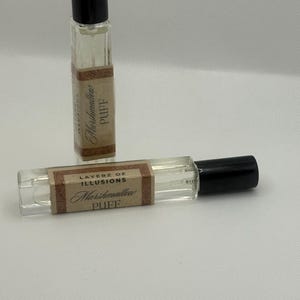 May include: Two small, clear glass perfume bottles with black caps. Each bottle has a brown label with the words "Layerz of Illusions Marshmallow Puff" printed in a decorative font. The bottles are on a white surface.