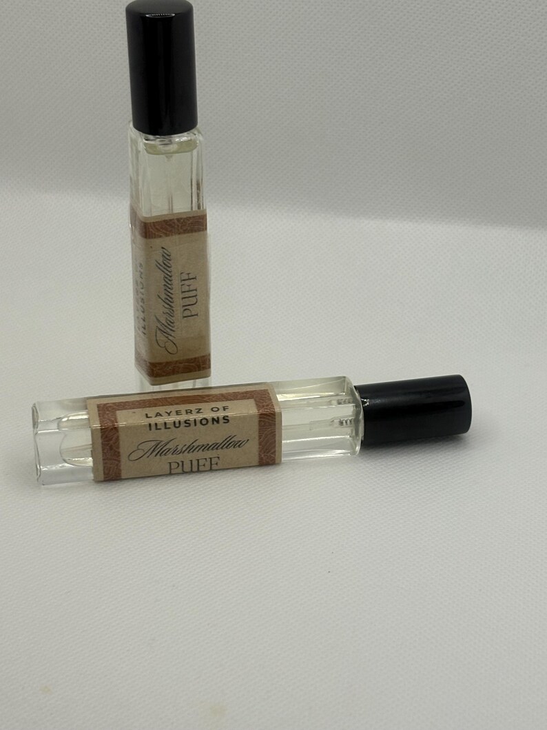 May include: Two small, clear glass perfume bottles with black caps. Each bottle has a brown label with the words "Layerz of Illusions Marshmallow Puff" printed in a decorative font. The bottles are on a white surface.