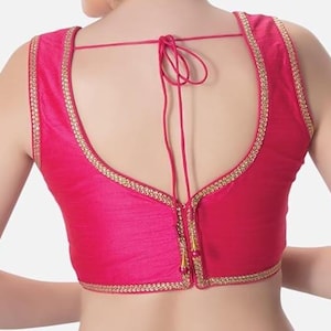 May include: A sleeveless, fuchsia-colored blouse with a U-shaped back. The edges are trimmed with a gold-colored, decorative border. The back features a drawstring closure and a decorative tassel.