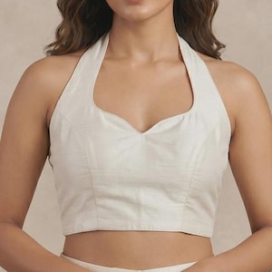 Puede incluir: Un top halter sin mangas de color blanco roto con escote corazón y dobladillo corto. La prenda está confeccionada con costuras estructuradas a lo largo del corpiño delantero, creando una silueta ajustada que se sitúa sobre la cintura.