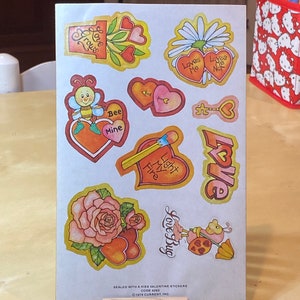 May include: A sheet of Valentine's Day stickers featuring various designs, including hearts, flowers, bees, and a ladybug. The stickers are printed on white paper and have a colorful, cartoonish style. The text on the sheet reads "Sealed with a Kiss Valentine Stickers, Code 4263, © 1979 Current, Inc."