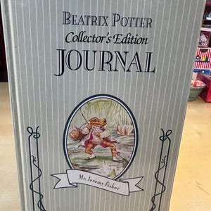 May include: A light gray journal with thin white vertical stripes. The cover features the text Beatrix Potter Collector's Edition JOURNAL and an oval illustration of Mr. Jeremy Fisher, a frog character holding a fishing rod and basket.