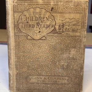 May include: Antique book titled "The Children's Third Reader" with a tan cover. The cover features a seashell design with the title in a decorative font. The publisher's name, Ginn & Company, is also visible. The book has a worn appearance.