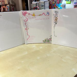 May include: Three white photo albums with decorative corner designs. One album has a red and green floral design, another has pink ribbons and text that reads "This is a Story About Me", and the third has a floral design.