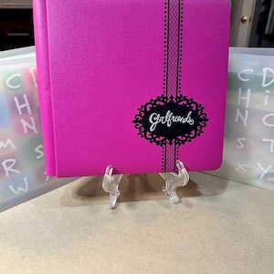 May include: A bright pink journal with the word "Girlfriends" in script, framed by a black decorative oval. A black lace design runs vertically down the journal. The journal is displayed on a clear stand, with alphabet letters in the background.