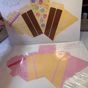 May include: Two flat, unfolded paperboard treat boxes. One is designed to resemble a slice of cake with chocolate cake layers, yellow frosting, and colorful candy decorations. The other is pink and yellow with a pink frosting design.
