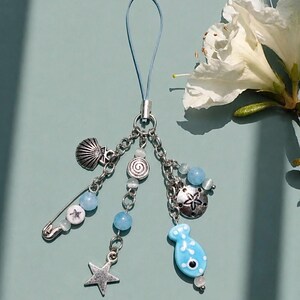 Può includere: Un ciondolo blu e argento con vari pendenti a tema oceano. Il ciondolo include una conchiglia, una stella e un pendente a forma di pesce blu. Il ciondolo è attaccato a un cordino blu, adatto per un portachiavi o un ciondolo per borsa.