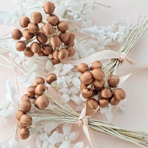 May include: Close-up of several bundles of decorative floral stems. Each bundle features clusters of small, round, bronze-colored buds. The stems are tied with a light pink ribbon and set against a backdrop of white floral accents on a soft pink surface.