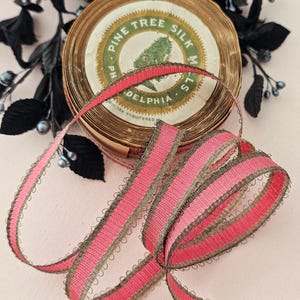 May include: A spool of vintage ribbon with a pink and gold design. The ribbon is made of silk and has a scalloped edge. The spool has a label that reads "Pine Tree Silk Mills, Philadelphia."