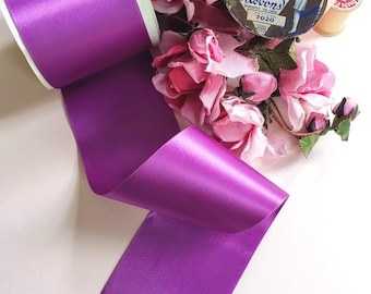 Puede incluir: Una bobina ancha de cinta de satén de color morado vibrante con un acabado liso y reflectante. La cinta está desenrollada y muestra un extremo cortado en zigzag. Cerca hay rosas artificiales de color rosa y una lata redonda vintage.