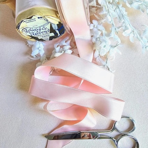May include: A spool of light pink taffeta ribbon with a vintage label reading "Reliable Ribbons." Silver scissors are positioned near the ribbon. White floral accents are in the background. The ribbon is unspooled and ready for use.