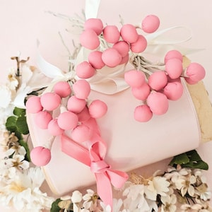May include: A soft pink, decorative arrangement featuring clusters of pink fabric berries, white ribbon, and a pink ribbon bow. The arrangement is accented with white flowers and greenery, creating a delicate and feminine aesthetic.