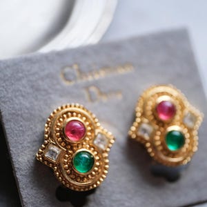 Op de afbeelding: Een paar goudkleurige clip-oorbellen met een textuurrijke, kralenrand. Elke oorbel is bezet met een ronde roze cabochon, een ronde groene cabochon en twee heldere, vierkant geslepen stenen, bevestigd op een grijze displaykaart.