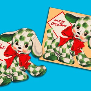 Puede incluir: Un perro de cuadros verdes y blancos sosteniendo una tarjeta que dice "Feliz Navidad". El perro lleva un lazo rojo y tiene una cara blanca con ojos negros.