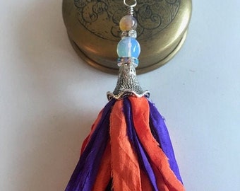 Puede incluir: Un borla de color morado y naranja con un abalorio de cuentas plateadas y blancas. La borla está hecha de tela y tiene un borde deshilachado.