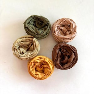 May include: Five circular bundles of frayed-edge silk ribbon arranged in a cluster. The colors include olive green, pale blush, golden yellow, tan, and deep chocolate brown. Each ribbon features a soft, wrinkled texture with visible loose threads.