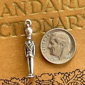 May include: A silver charm shaped like a toy soldier. The charm is approximately the size of a ten pence piece. The soldier is wearing a hat and a uniform with a belt and a sword.
