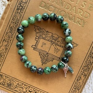 May include: A green and black gemstone bracelet with a silver key charm. The bracelet is on top of an old book with the word "STANDARD DICTIONARY" printed on the cover.