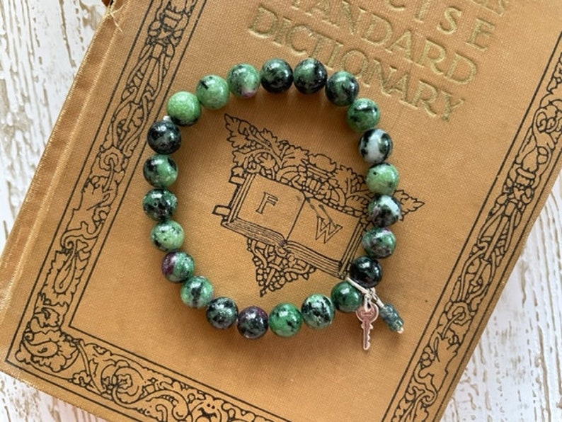 May include: A green and black gemstone bracelet with a silver key charm. The bracelet is on top of an old book with the word "STANDARD DICTIONARY" printed on the cover.