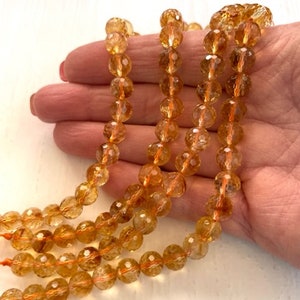 May include: A strand of golden citrine beads, faceted and translucent, with a warm, honey-like color. The beads are strung on a clear elastic cord and are approximately 8mm in diameter.