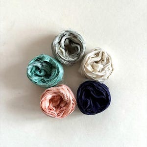 May include: Five spools of fabric ribbon in different colors: light blue, dark blue, pink, white, and gray. The spools are arranged in a circle on a white background.