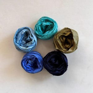 May include: An overhead shot displays five fabric rolls in a circular arrangement. The rolls are in shades of blue, teal, and olive green. The fabric appears to be textured and possibly silk or a similar material, ideal for crafting or decorative purposes.