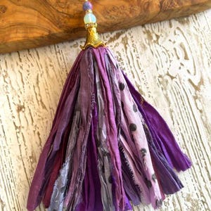 May include: A decorative tassel made of multiple strips of purple and lavender fabric, some featuring a dark polka dot pattern. The top is secured with a gold-toned metal cap, a small clear bead, and a purple bead, finished with a small wire loop.