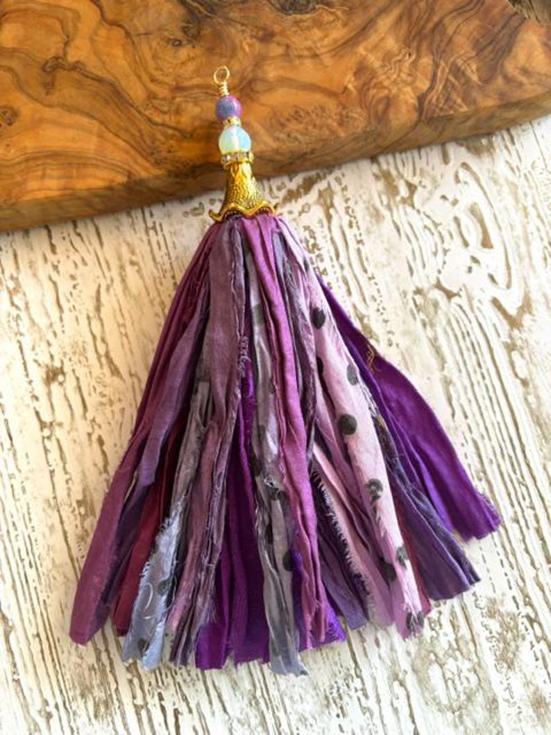 May include: A decorative tassel made of multiple strips of purple and lavender fabric, some featuring a dark polka dot pattern. The top is secured with a gold-toned metal cap, a small clear bead, and a purple bead, finished with a small wire loop.