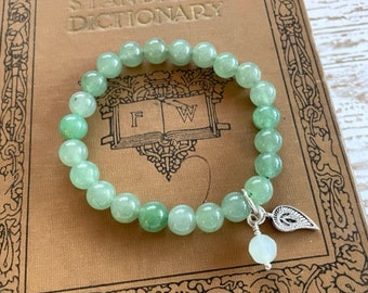 May include: A green gemstone bracelet with a silver charm in the shape of a leaf. The bracelet is made of round beads and is strung on a clear elastic cord.