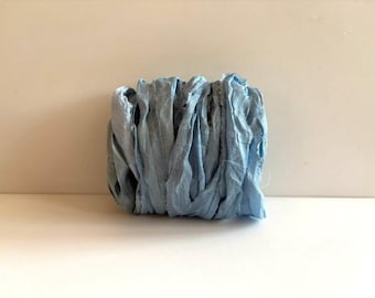May include: A bundle of light blue silk ribbon with a crinkled, textured surface and frayed edges. The ribbon is wound into a compact, cylindrical shape, showcasing a soft, matte finish and consistent pale blue color throughout the material.