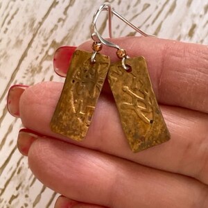 May include: A pair of gold-toned earrings with a rectangular shape and a textured surface. The earrings are attached to silver hooks with copper beads.