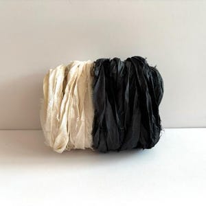 Könnte beinhalten: Zwei Spulen mit Seidenband, eine weiße und eine schwarze. Das Band ist ausgefranst und hat einen rustikalen Look.