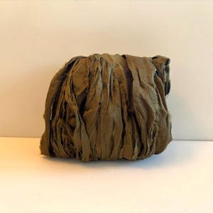 May include: A bundle of olive green silk ribbon featuring a raw, frayed edge texture. The fabric is gathered into a dense, rounded cluster with visible vertical folds and creases throughout the material, showcasing a matte, earthy color tone.