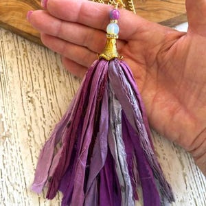 May include: A tassel pendant featuring multiple strips of fabric in various shades of purple, lavender, and grey. The tassel is topped with a gold-toned metal cap, a white bead, a small purple bead, and a gold-toned chain necklace.