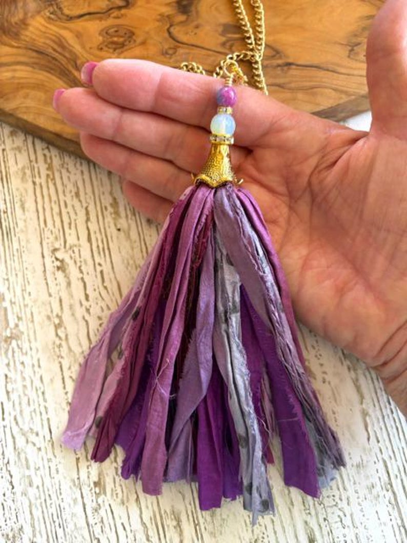 May include: A tassel pendant featuring multiple strips of fabric in various shades of purple, lavender, and grey. The tassel is topped with a gold-toned metal cap, a white bead, a small purple bead, and a gold-toned chain necklace.