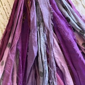May include: A collection of fabric strips in various shades of purple, lavender, and light gray. The strips feature frayed edges and a mix of solid colors and patterns, including some with small dark polka dots, hanging vertically in a dense cluster.